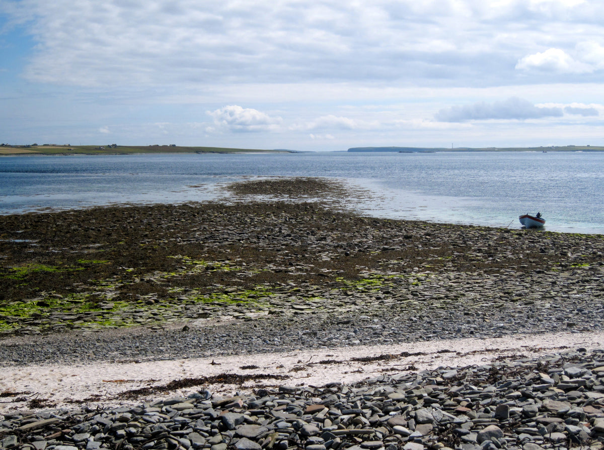 A stony beach in the foreground looking out to sea with Westray and Papay in background