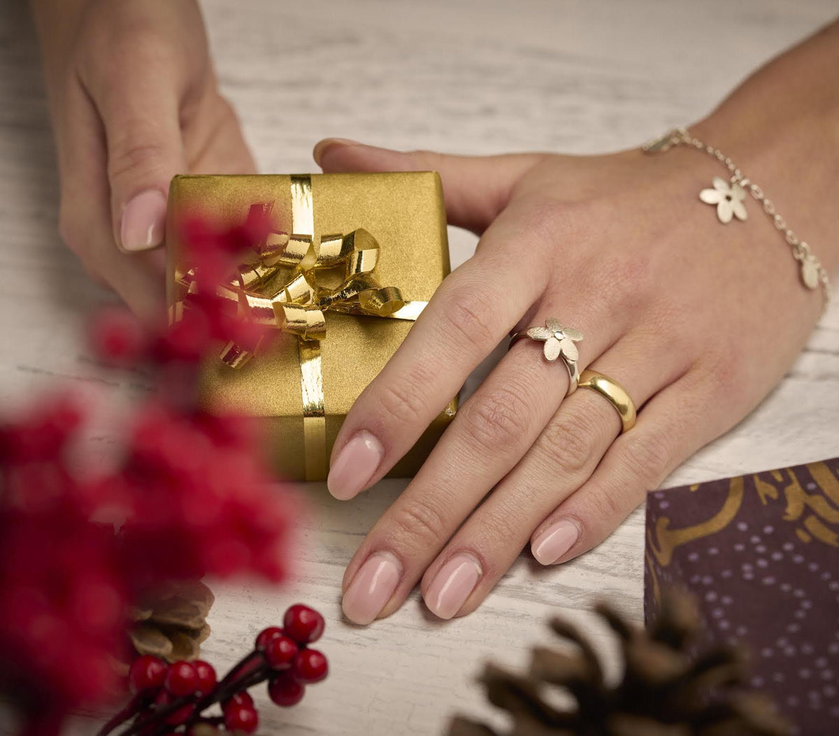 A pair of hands holding a small, square, gold-colored gift box with a decorative ribbon, surrounded by festive decorations like red berries and pine cones.