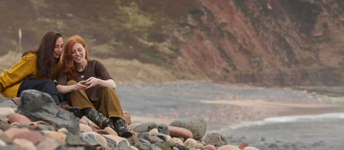 Two women are sitting on the stones at Rackwick Bay, looking a pebbles whilst modelling a silver necklace
