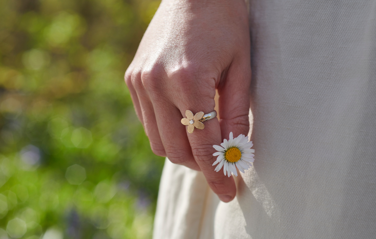 Hand wearing a gold ring with a floral design, holding a white daisy against a blurred natural background.