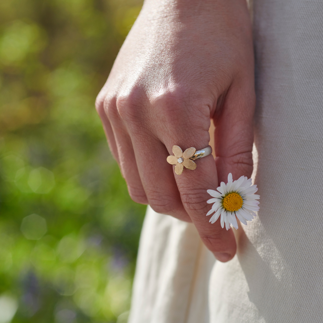 Hand wearing a gold flower ring with a daisy held against a blurred green background