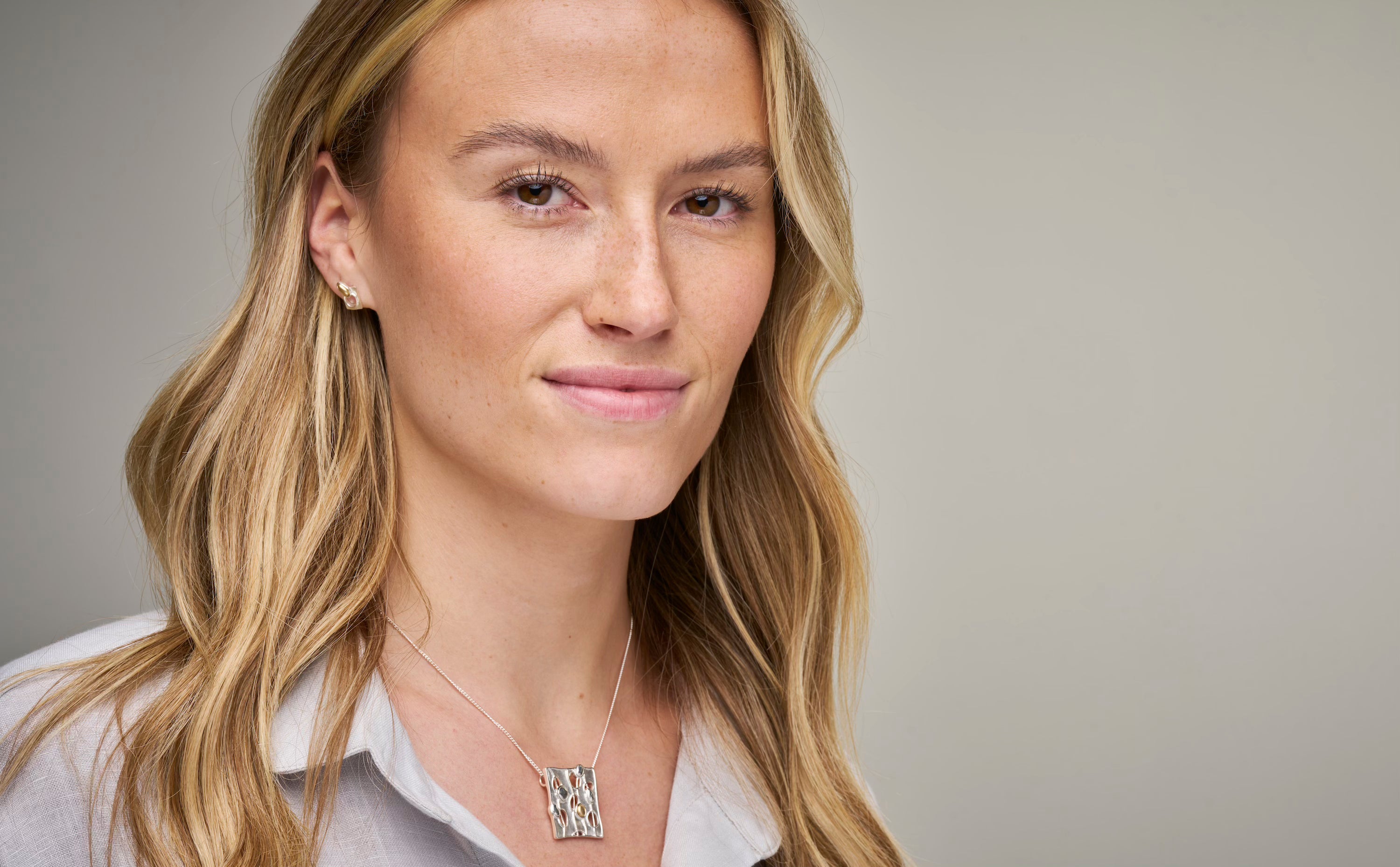 Woman facing camera in grey top with square necklace and studded earring