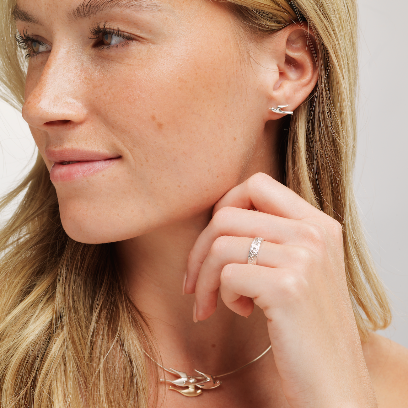 Closeup of a woman wearing a silver birdland pendant on a chain necklace with matching stud earring and rysa ring