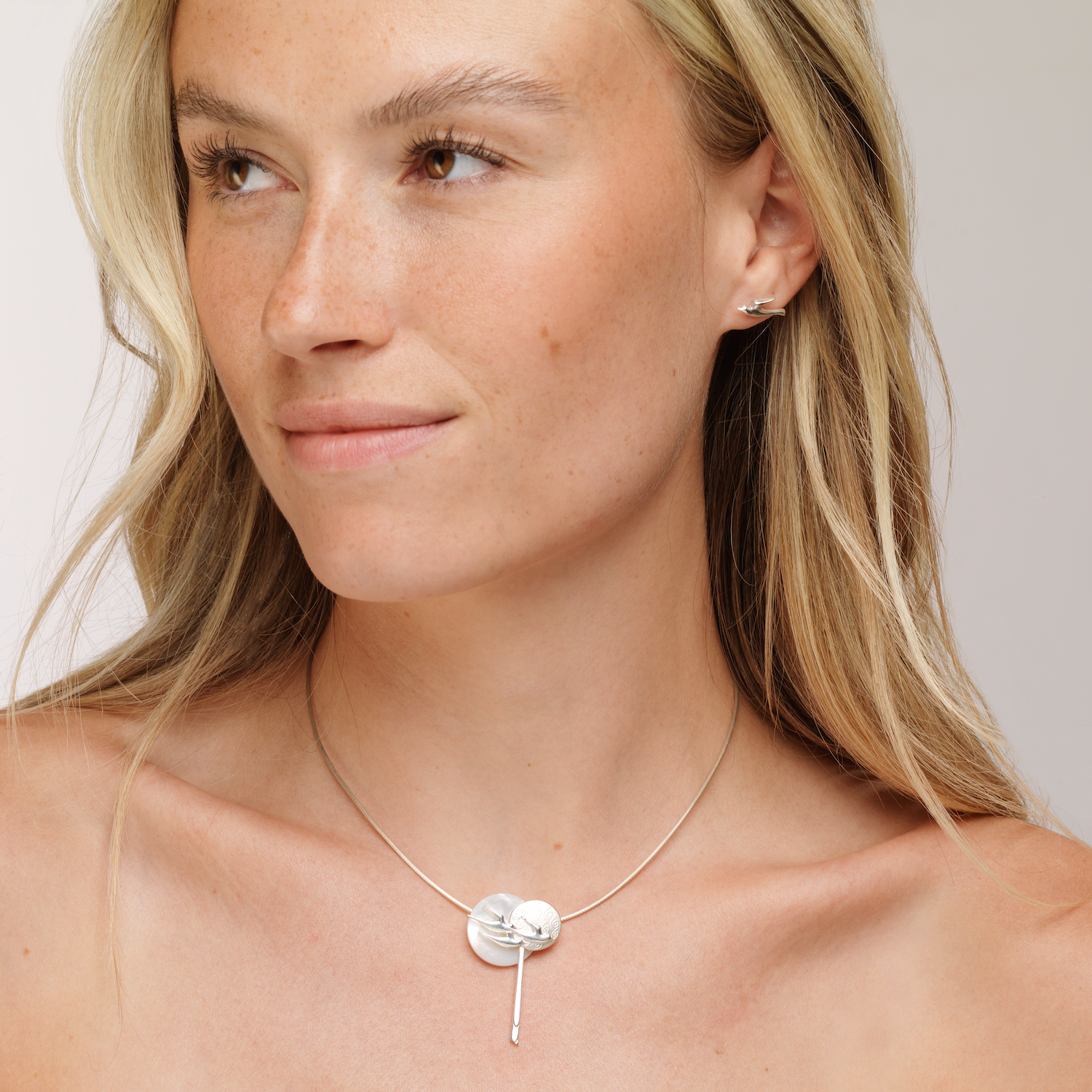 A woman wearing a silver birdland pendant on a chain necklace with matching stud earring