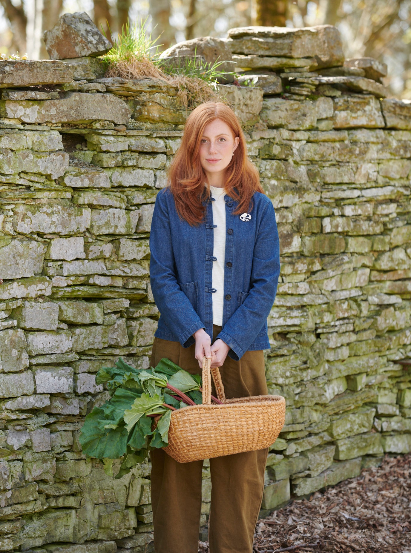 A woman is standing in front of a stone wall holding a basket of rhubarb and modelling a brooch