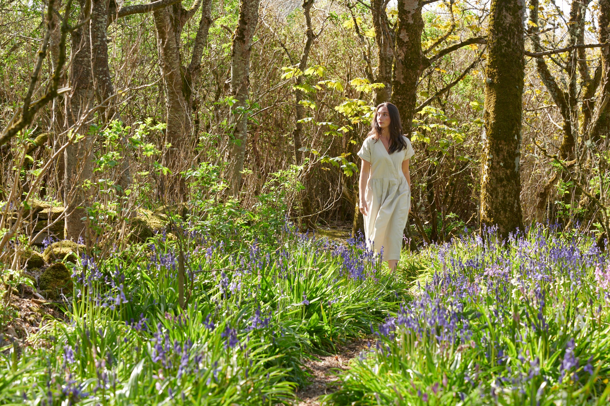 A woman in white dress is walking through a wood with bluebells